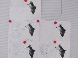 Giraffensprache sechs schwarz weiße arbeitsblätter mit wolfsillustrationen sind an eine tafel gepinnt. jedes blatt enthält handgeschriebenen deutschen text in und um sprechblasen, die an den wolf gerichtet sind. vier rote stecknadeln halten die blätter an ihrem platz.