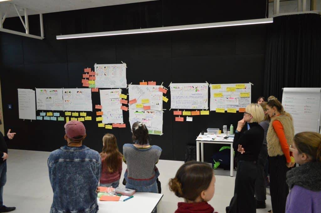 eine gruppe von personen steht in einem raum vor einer schwarzen wand mit großen papieren, tabellen und bunten haftnotizen, die in reihen angeordnet sind, als ob sie an einem workshop oder einer gemeinsamen brainstorming sitzung teilnehmen würden. eine gruppe von personen steht in einem raum vor einer schwarzen wand mit großen papieren, tabellen und bunten haftnotizen, die in reihen angeordnet sind, als ob sie an einem workshop oder einer gemeinsamen brainstorming sitzung teilnehmen würden.