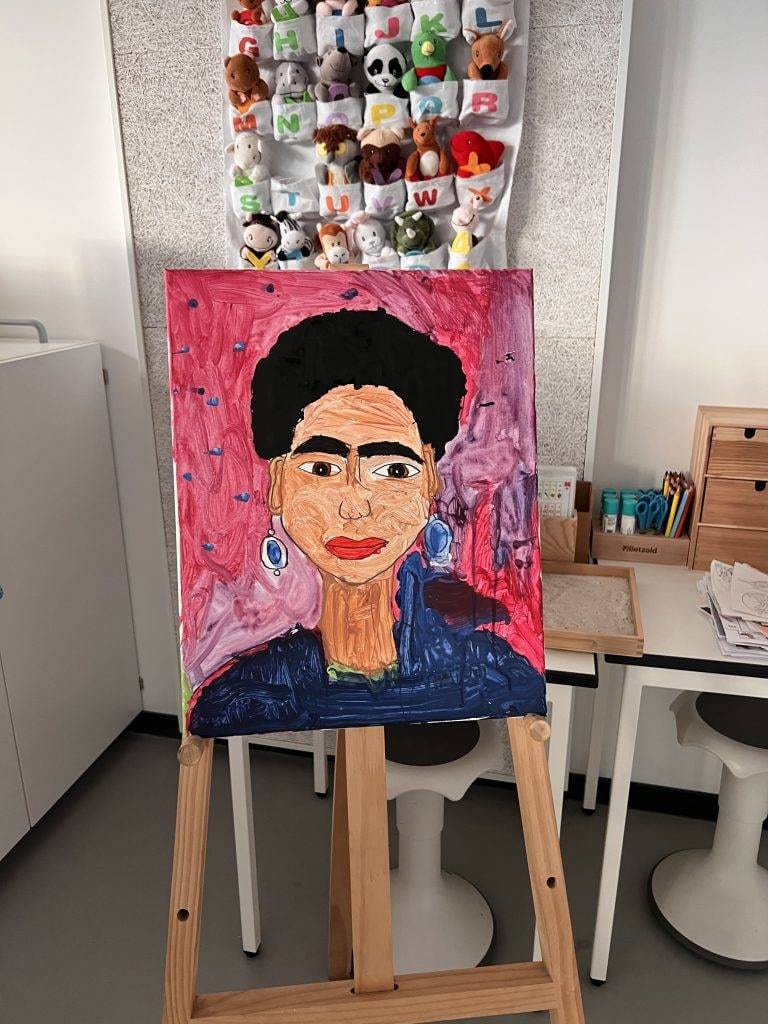 Frida-Kahlo-Projekts ein farbenfrohes gemälde einer frau mit ohrringen, dunklem haar und einem blauen oberteil ist auf einer staffelei in einem raum mit spielzeug, kunstzubehör und kindermöbeln ausgestellt. der hintergrund des bildes ist rosa.