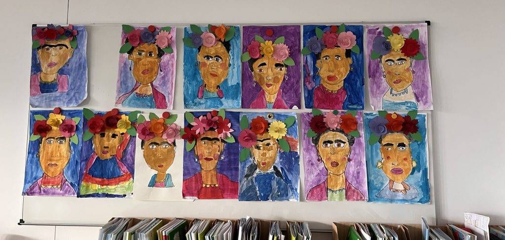 Frida-Kahlo-Projekts an einer wand im klassenzimmer hängen farbenfrohe porträts von frida kahlo, die sich durch helle hintergründe, markante gesichtszüge und 3d papierblumen als kopfschmuck auszeichnen. in einem regal unterhalb der kunstwerke sind reihen von büchern zu sehen.
