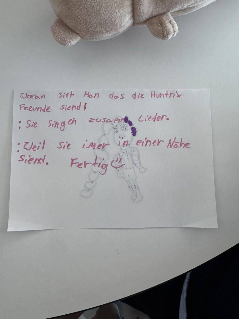 eine handschriftliche notiz in deutscher sprache auf weißem papier, verziert mit einer schwachen skizze von zwei personen. am oberen und unteren rand des bildes sind ein plüschtier und ein teil eines runden tisches zu sehen.