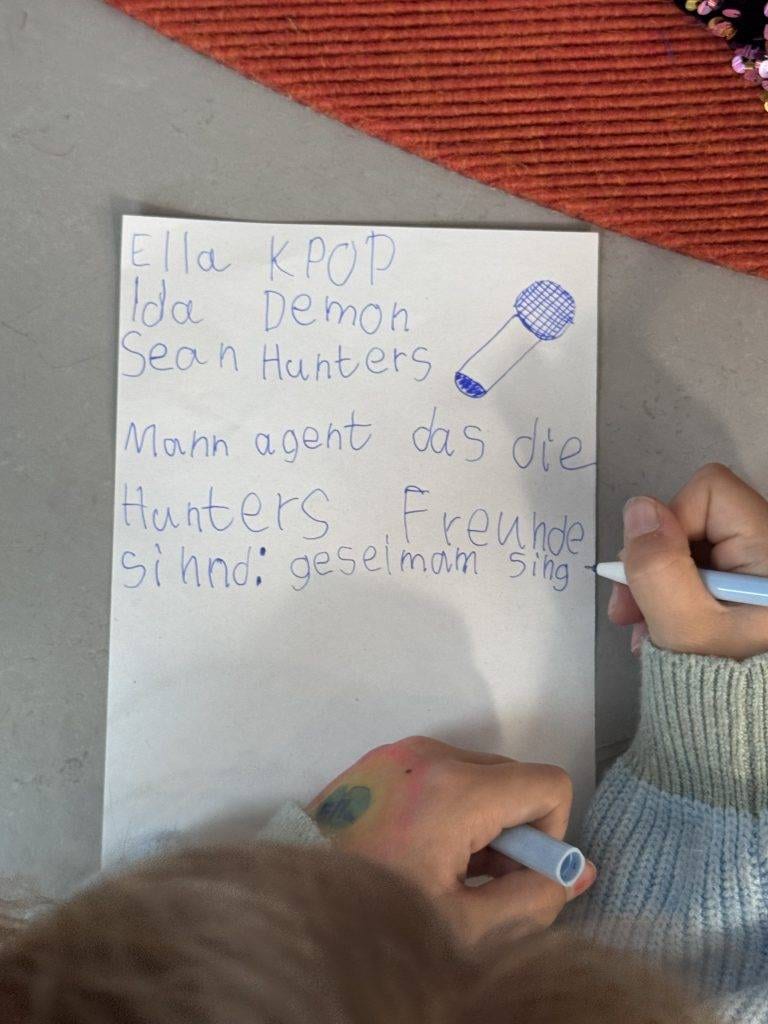 ein kind schreibt mit blauem filzstift auf ein blatt papier. auf dem zettel, der in deutscher sprache verfasst ist, stehen namen, ein kritzelndes mikrofon und die worte freunde sind: gemeinsam singen. man sieht die hände des kindes, die den marker halten.
