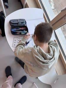 Klasse ¾, immer donnerstags ein kind sitzt an einem weißen schreibtisch und zeichnet mit buntstiften. auf dem tisch liegen ein offenes federmäppchen und papiere mit zeichnungen. das kind trägt einen hellgrauen kapuzenpullover und arbeitet in der nähe eines großen fensters, in dessen nähe eine weitere person steht.