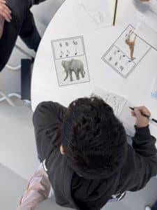 Klasse ¾, immer donnerstags ein kind sitzt an einem runden weißen tisch und malt ein tier auf papier. auf dem tisch liegen tierkarten mit musiknoten und abbildungen eines elefanten, eines dinosauriers und einer giraffe. andere kinder sind teilweise sichtbar.