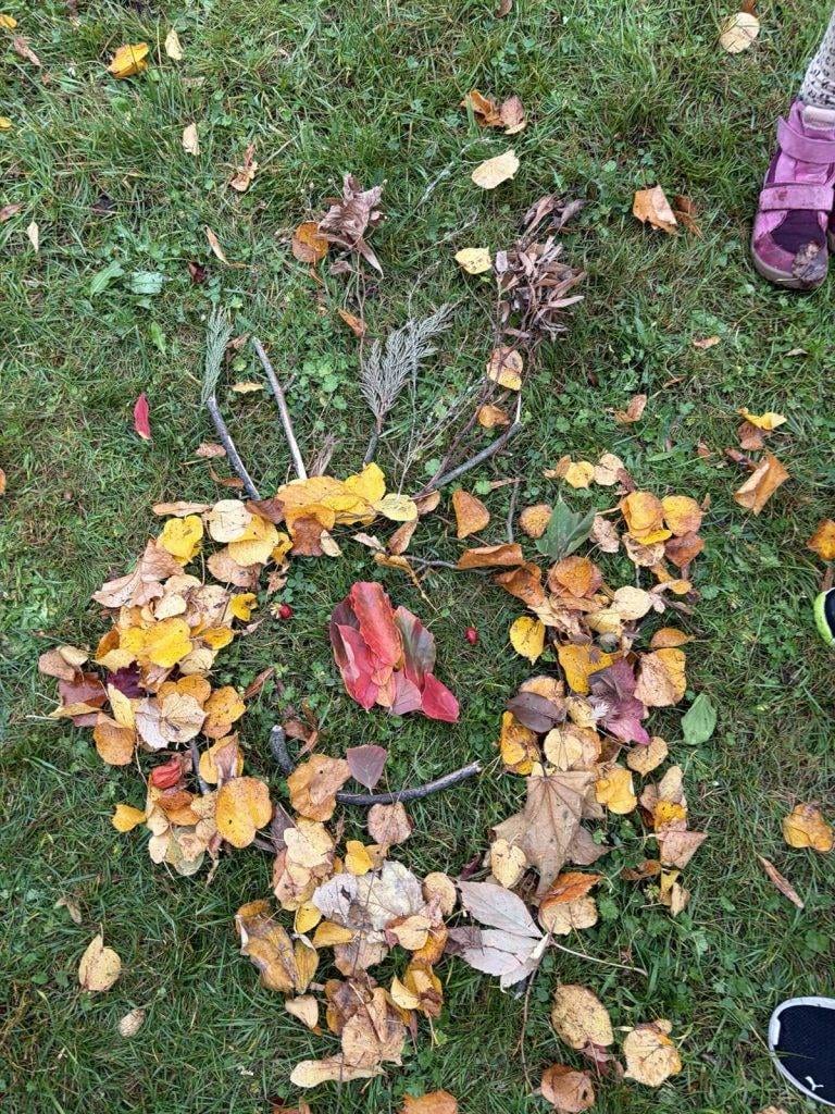 ein arrangement aus herbstblättern und stöcken auf gras bildet die form eines hasengesichts mit zwei ohren an der spitze. an den rändern des bildes sind teilweise zwei schuhe zu sehen.
