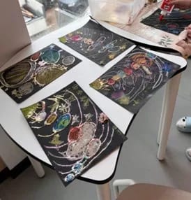 vier bunte buntstift und glitzerzeichnungen des sonnensystems auf schwarzem papier sind auf einem kleinen weißen tisch in der nähe eines fensters ausgestellt. in der oberen rechten ecke ist eine hand mit bastelmaterial zu sehen.
